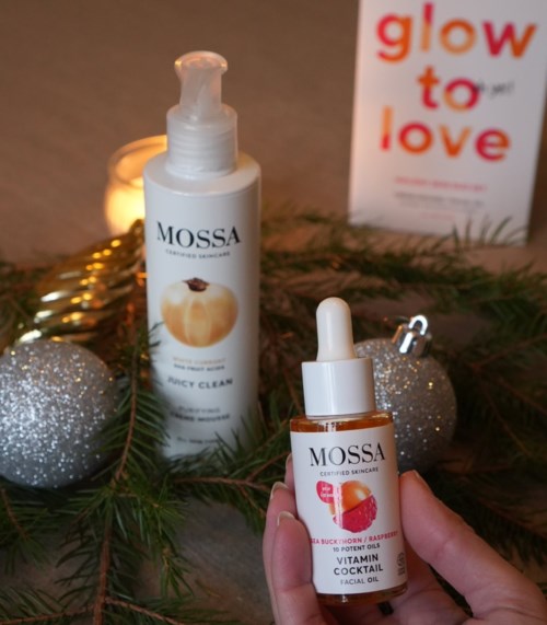 Een hand houdt een bruine fles met pipet, omgeven door feestdecoraties en een ander huidverzorgingsproduct. Een bord zegt "glow to love."