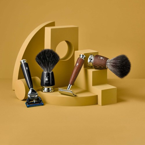 Kokoelma partavälineitä, kuten vapaaporakone, parta-ateria, safety razor ja hoitoharja beigen geometrisessä taustassa.