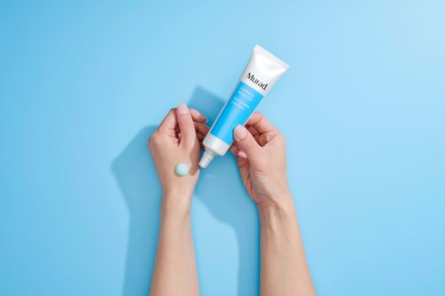 Handen houden een tube gezichtscrème vast op een blauwe achtergrond, met crème op de hand. De tube is wit en blauw.