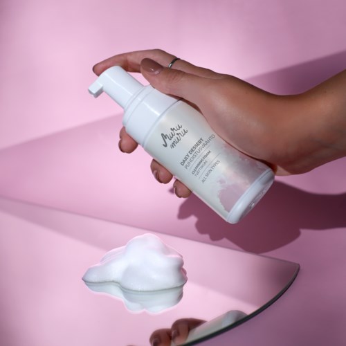 A hand holds a white pump bottle labeled 'Baby Organic,' with foam on a mirror surface. Pink background.