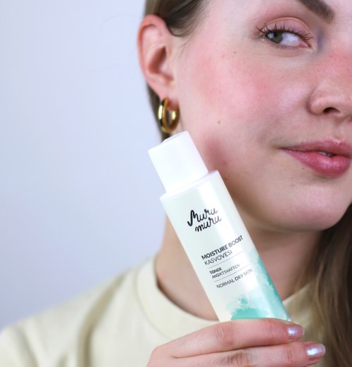 A woman with fair skin and gold hoop earrings holds a white bottle of skincare product near her face.