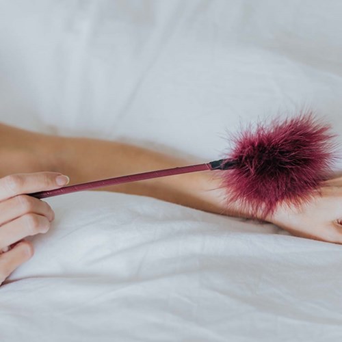 Eine Person hält einen pinken Zauberstab mit einer flauschigen pinken Spitze, ruht auf einem weißen Bett.