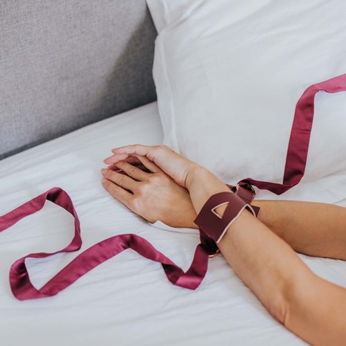 Die Hände einer Person liegen auf einem Bett, mit einem Band oder Riemen um das Handgelenk, und weiße Kissen sind sichtbar.