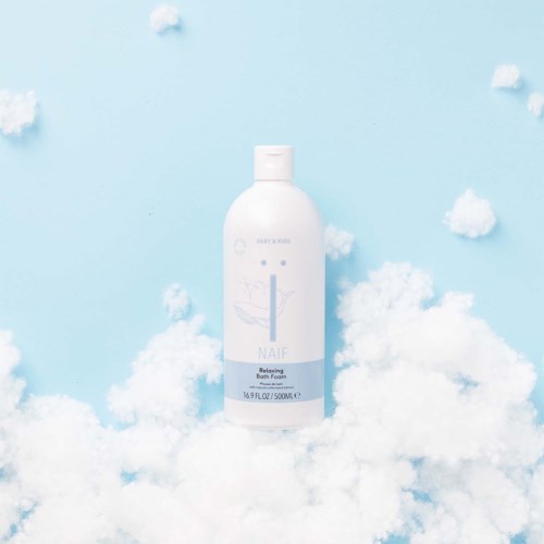 Eine weiße Flasche Naïf Relaxing Bath Foam umgeben von Wolken und Baumwolle. Heller blauer Hintergrund. Minimalistisches Design.