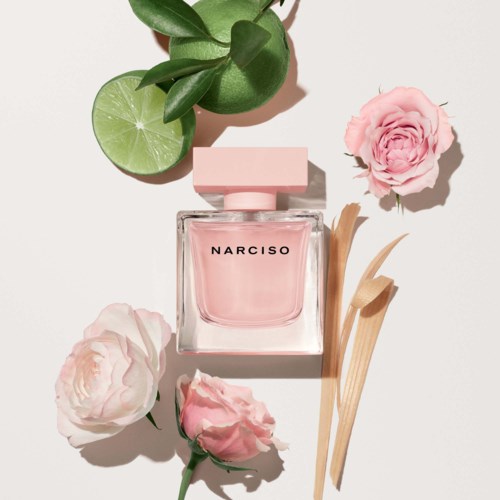 Een flacon met 'Narciso' parfum, omgeven door roze rozen, groene limoen en beige houten stokjes op een lichte achtergrond.