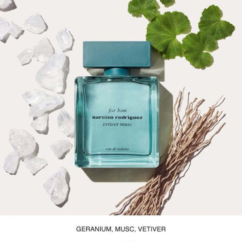 Een parfumfles met groene bladeren, takjes en witte stenen eromheen. De fles is blauw en voor hem gelabeld.