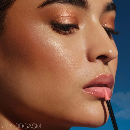 A woman applying gloss on her lips, with shiny makeup and closed eyes, against a blue background.