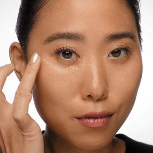 A woman touches her face with one hand, showing clear skin and natural makeup, against a plain white background.