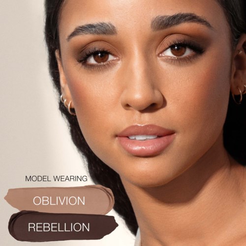 Een vrouw met een egale huid en bruine ogen, toont een subtiele glimlach. Make-up omvat oogschaduw, lippenstift en wenkbrauwen.