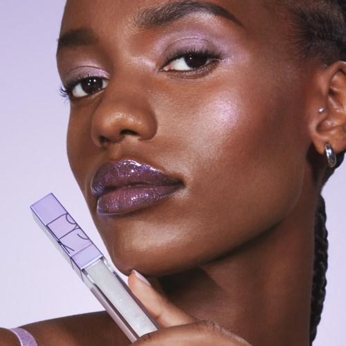 A woman with shiny purple lipstick holds a lip gloss tube, showing her glossy lips and smooth skin.