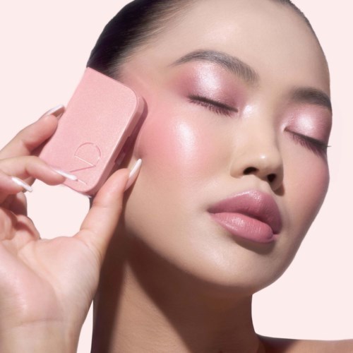 Eine Frau mit geschlossenen Augen benutzt einen rosa Make-up-Schwamm auf ihrer Wange und zeigt einen klaren, strahlenden Teint.