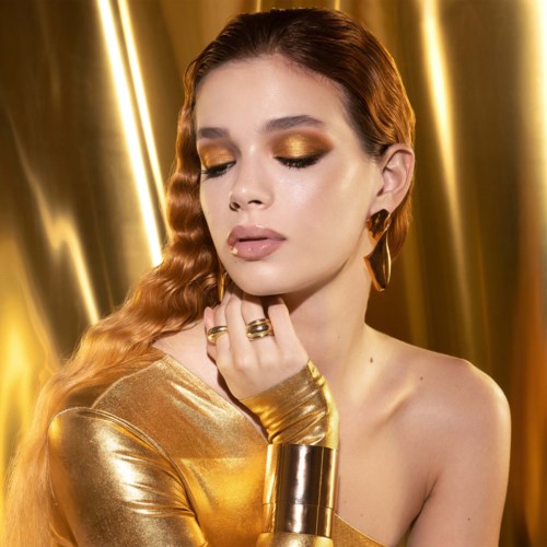 Eine Frau mit welligem roten Haar, goldigem Make-up, goldenem Kleid und Goldschmuck steht vor einem glänzenden goldenen Hintergrund.