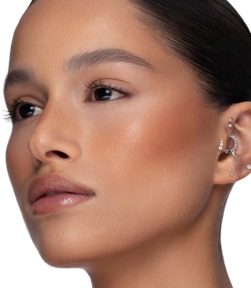 Eine Frau mit glatter Haut und dunklem Haar, trägt mehrere Ohrringe und minimalistisches Make-up, wirkt ruhig und selbstbewusst.
