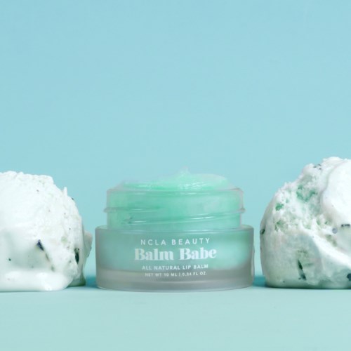 A small green jar labeled 'Balmy Babe All Natural Lip Balm' with two white fluffy objects on a blue background.