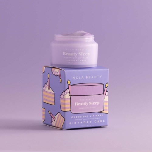 Ein Behälter mit Beauty Sleep Creme auf einer Schachtel mit Geburtstagskuchen und Kerzen, vor violem Hintergrund.