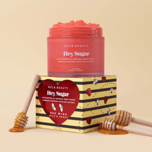Ein rosa Körperschleifglaskrug steht auf einer bunten Schachtel mit Bienen- und Honigwaben-Design, umgeben von Honighörnchen.