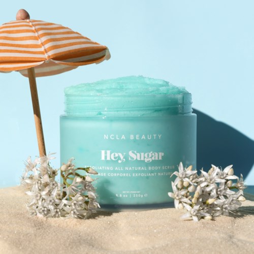 Eine hellblaue Dose mit Körperpeeling, ein Strandschirm und weiße Blumen im Sonnenlicht.