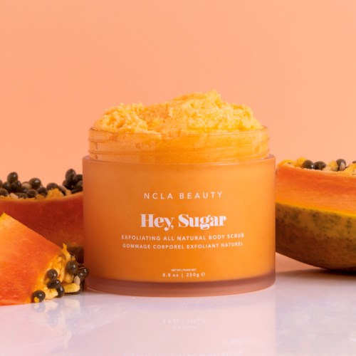 A yellow body scrub container surrounded by papaya slices and black seeds on a white surface.