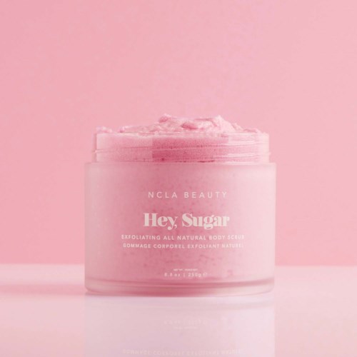Ein rosa Behälter mit Körperpeeling mit schaumiger Textur, gekennzeichnet 'Hey, Sugar' für Hautpeeling.