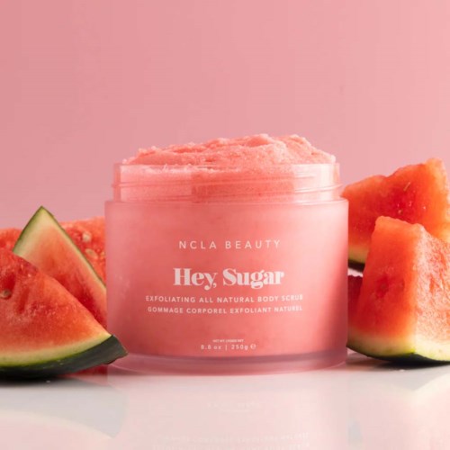 Ein pinker Body-Scrub-Topf mit Wassermelonenstücken um ihn herum, auf rosa Hintergrund.