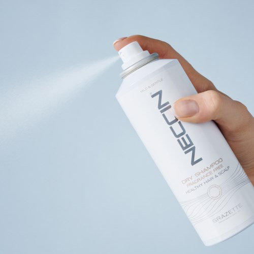 Een hand spuit een wit blikje NeoCos droogshampoo voor haar en hoofdhuid. De achtergrond is lichtblauw.