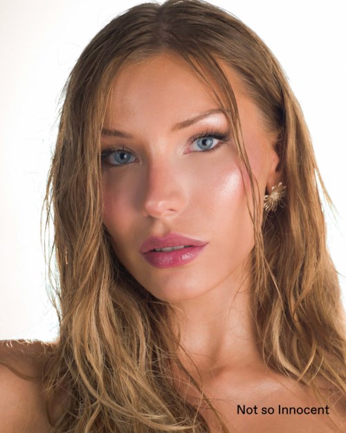 A woman with long wavy blonde hair, blue eyes, light makeup, and a slight smile. She wears earrings and a sleeveless top.