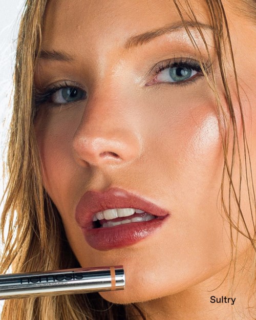 A woman with wet hair, blue eyes, and glossy lips holds a lipstick near her mouth, looking at the camera.