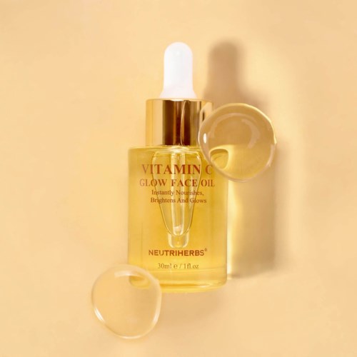 A bottle of Vitamin C glow face oil with a dropper, placed on a cream-colored background, with two gel-like drops nearby.