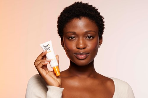 Een vrouw met kort kroes haar gebruikt een huidverzorgingsproduct genaamd NIP+FAB en houdt een tube met oranje dop.