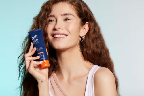 Een glimlachende jonge vrouw met lang krullend haar houdt een tube NIP+FAB huidverzorgingsproduct.