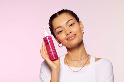 Een vrouw houdt een roze fles met dispenser, glimlacht zachtjes voor een lichtroze achtergrond.