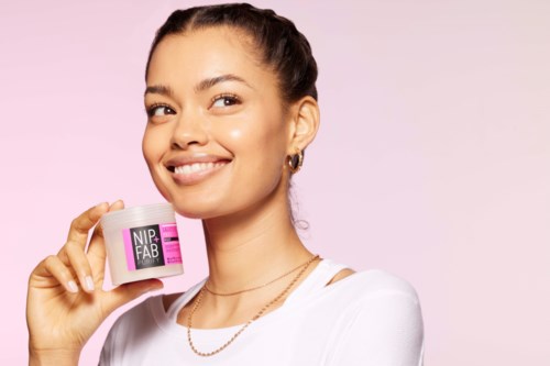 Een glimlachende vrouw houdt een kleine roze container vast, promoot een schoonheids- of huidverzorgingsproduct tegen een lichtroze achtergrond.