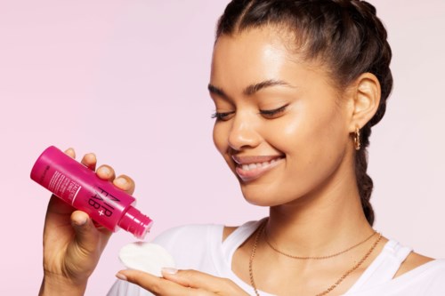 Een lachende vrouw met gevlochten haar giet shampoo op haar hand tegen een roze achtergrond.