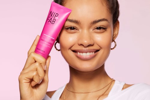 Een lachende vrouw houdt een roze tube huidverzorgingsproduct genaamd NIP+FAB vast tegen een roze achtergrond.
