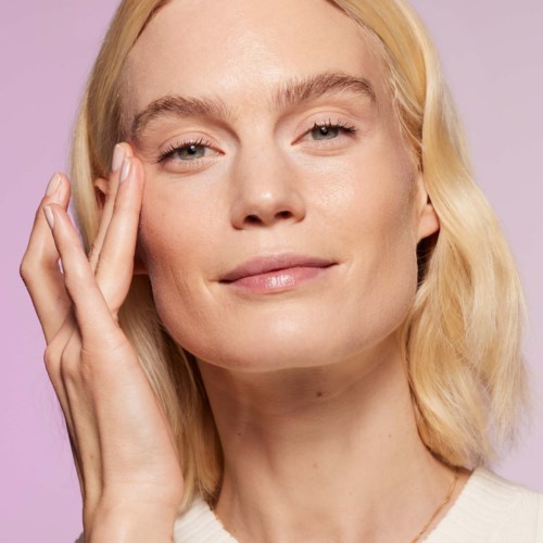 Een vrouw met blond haar raakt voorzichtig haar gezicht aan en glimlacht zachtjes tegen een lavendelkleurige achtergrond.