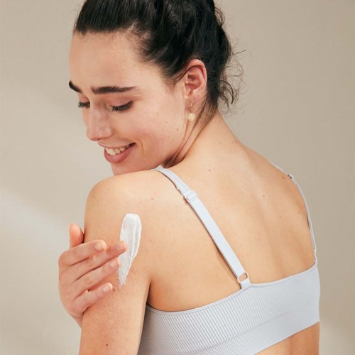 Een vrouw brengt lotion aan op haar schouder met een vriendelijke glimlach.