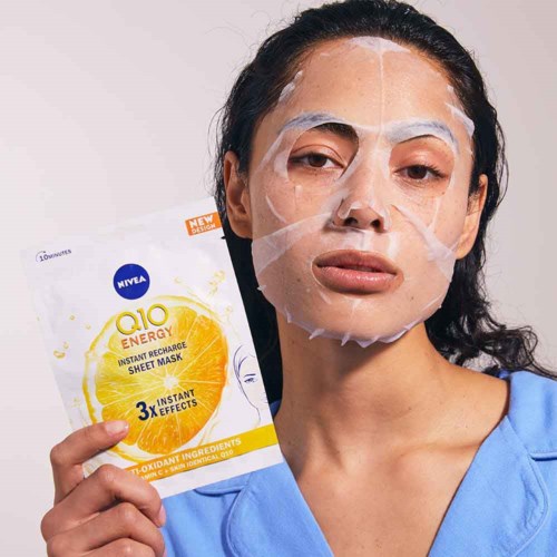 Een vrouw met een gezichtsmasker, houdt een Nivea Q10 energiemasker-verpakking, glimlacht zacht.