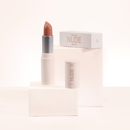 A beige lipstick stands on a white block with its cap beside, in front of a box labeled "NUDE BEAUTY."
