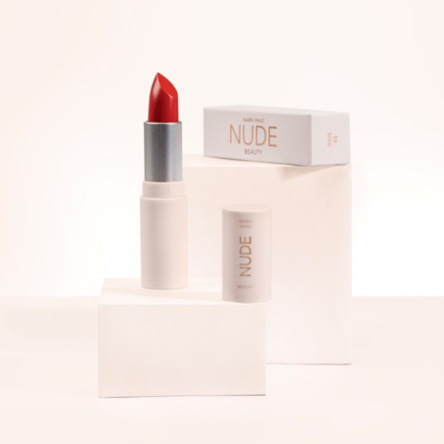 A red lipstick with white and gray packaging, placed on white blocks, with a box labeled 'NUDE BEAUTY' in the background.