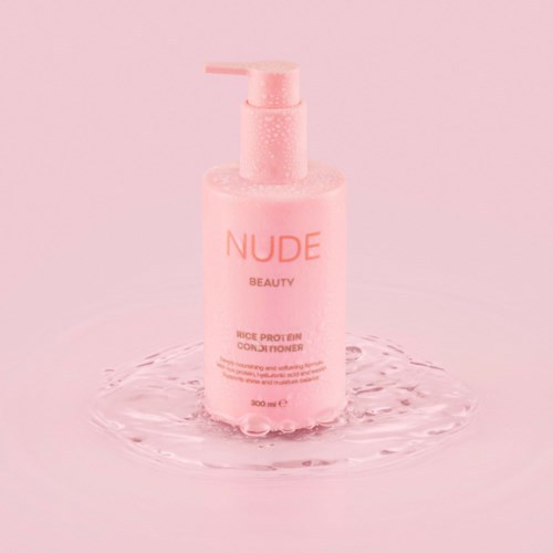 Ljusrosa flaska med etiketten NUDE BEAUTY och vatten runt den på en rosa bakgrund.