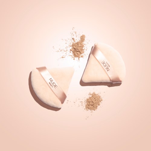 Two makeup sponges labeled 'NUDE' with powder spilled around them on a light background.
