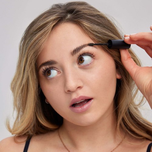 Een vrouw brengt mascara op haar wimpers aan, kijkt omhoog met een geïnteresseerd uitdrukking.