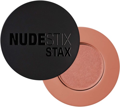 Een zwarte ronde container met "NUDESTIX STAX" erop, gedeeltelijk bedekt door roze blush of oogschaduw.