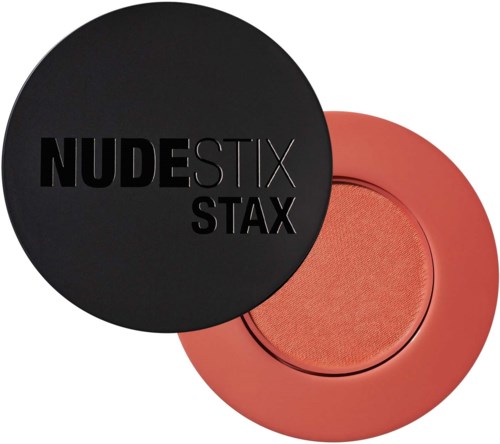 Ein runder schwarzer Behälter mit den Worten "NUDESTIX STAX" darauf, teilweise bedeckt von einem korallenfarbenen Makeup-Produkt.