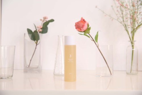 Een beige fles staat in het midden op een witte oppervlakte, omgeven door verschillende heldere glazen vazen met roze en witte bloemen.