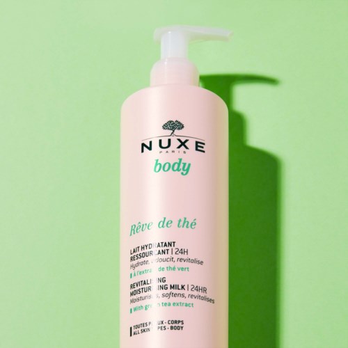 A pink bottle of Nuxe body lotion with a pump, green and black text, and a shadow on a green background.