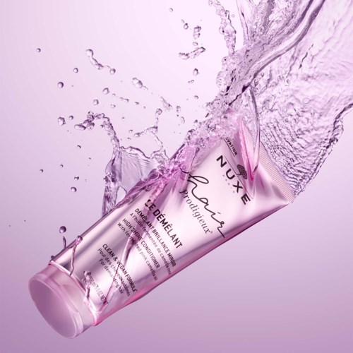 Een roze tube met 'NUXE' erop wordt gespoten met water en staat tegen een zachte roze achtergrond.