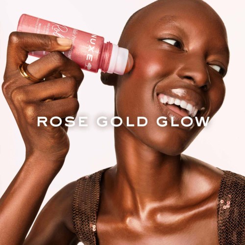 A smiling woman with dark skin uses a pink beauty product on her face. The words Rose Gold Glow are written.