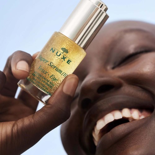 A person laughs happily while holding a small bottle of Nuxe skincare product near their face.