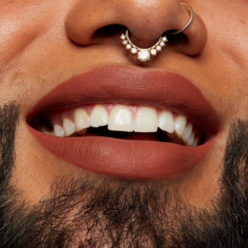 Close-up of a person with a nose piercing and an open smile showing teeth and lip makeup.
